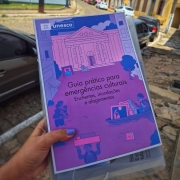 Livro com o t&iacute;tulo: &ldquo;Guia pr&aacute;tico para emerg&ecirc;ncias culturais: enchentes, inunda&ccedil;&otilde;es e alagamentos&rdquo;. Ele &eacute; segurado pelas m&atilde;os de uma mulher. O livro possui capa com cores em v&aacute;rios tons de roxo e rosa, demonstrando uma situa&ccedil;&atilde;o de inunda&ccedil;&atilde;o em uma &aacute;rea com pr&eacute;dios. Ao fundo a paisagem &eacute; de uma rua com casas antigas e carros estacionados do lado direito.